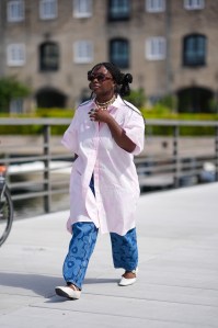 COPENHAGEN, DENMARK - AUGUST 8: A guest wears dark brown sunglasses, gold earrings, white and gold layered choker necklaces, silver rings, light pink button up short sleeve pattern shirt, navy blue light blue floral denim jean pants, white ballerina flats leather shoes, outside Marimekko, during the Copenhagen Fashion Week Spring/Summer 2024-2025 on August 8, 2024 in Copenhagen, Denmark. (Photo by Edward Berthelot/Getty Images)